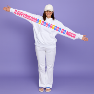 Woman wearing a white unisex hoodie with a colorful “I LOVE YOU SOOOOO MUCH” graphic printed across the sleeves and chest, standing with arms wide open.