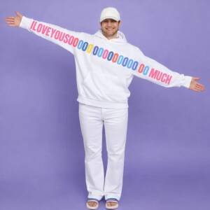 Man wearing a white unisex hoodie with a colorful “I LOVE YOU SOOOOO MUCH” graphic printed across the sleeves and chest, standing with arms wide open.