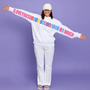 Woman wearing a white unisex hoodie with a colorful “I LOVE YOU SOOOOO MUCH” graphic printed across the sleeves and chest, standing with arms wide open.