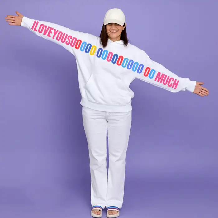 Woman wearing a white unisex hoodie with a colorful “I LOVE YOU SOOOOO MUCH” graphic printed across the sleeves and chest, standing with arms wide open.